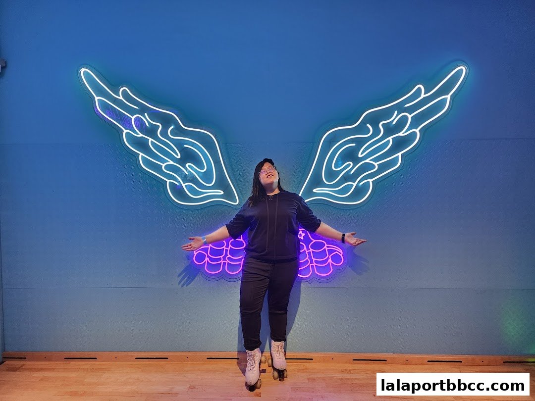 A person in roller skates poses with outstretched arms in front of a neon wing art installation.