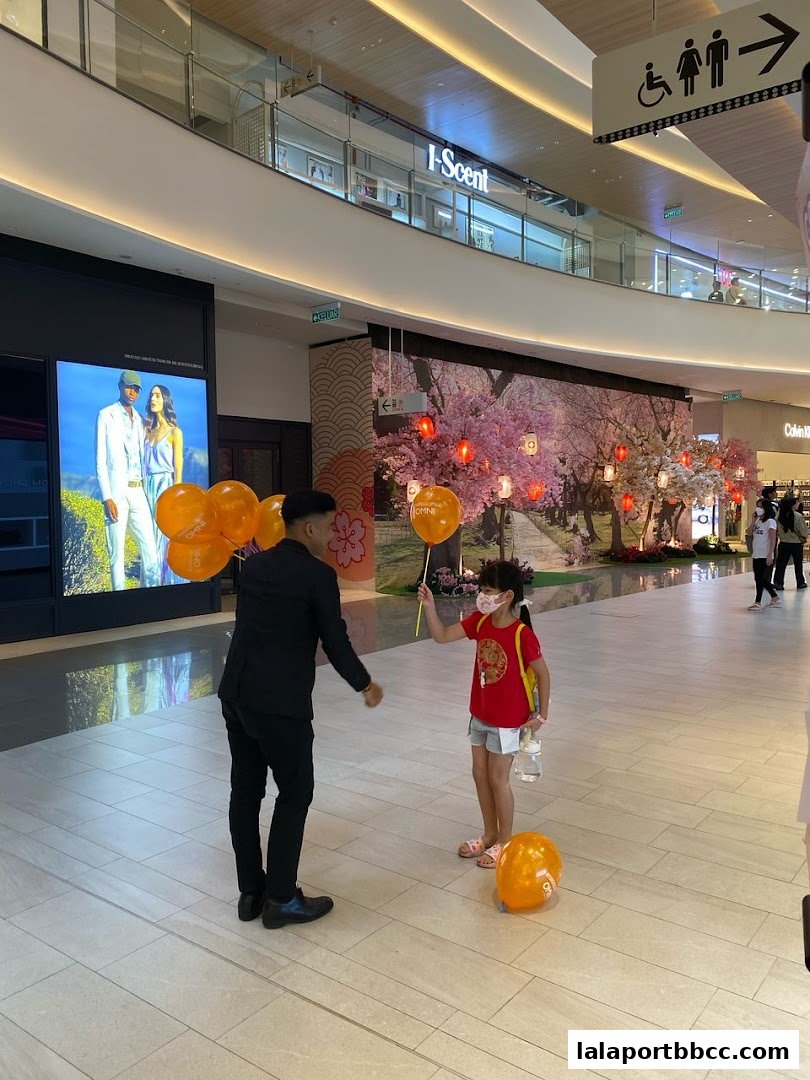 A staff member hands a balloon to a child in a mall with a decorative cherry blossom display.
