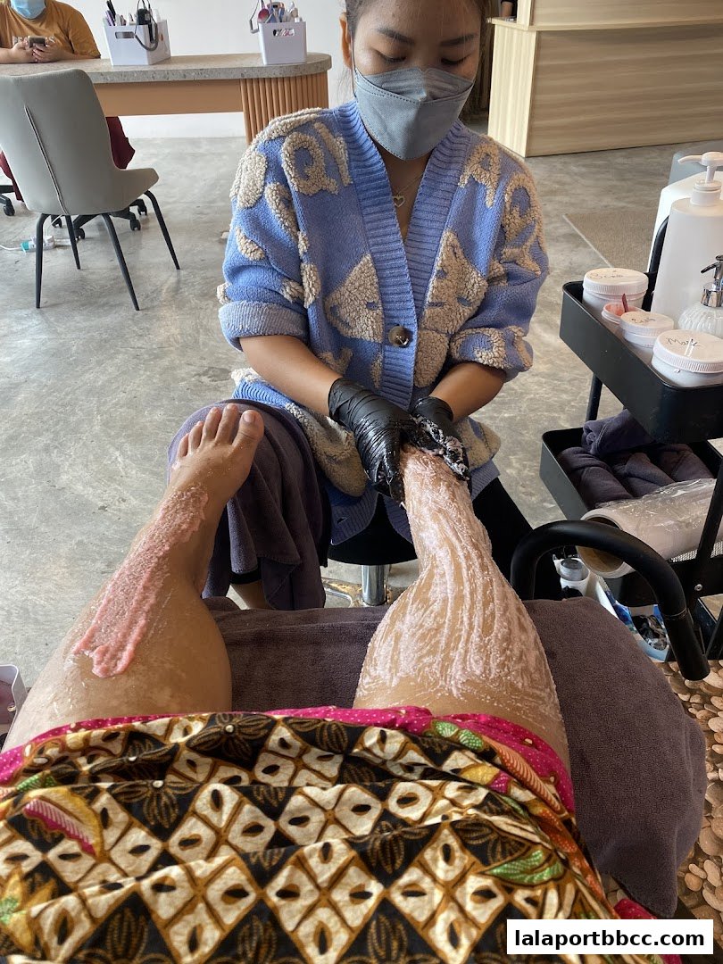 A nail technician performs a pedicure on a client's foot with a pink scrub.