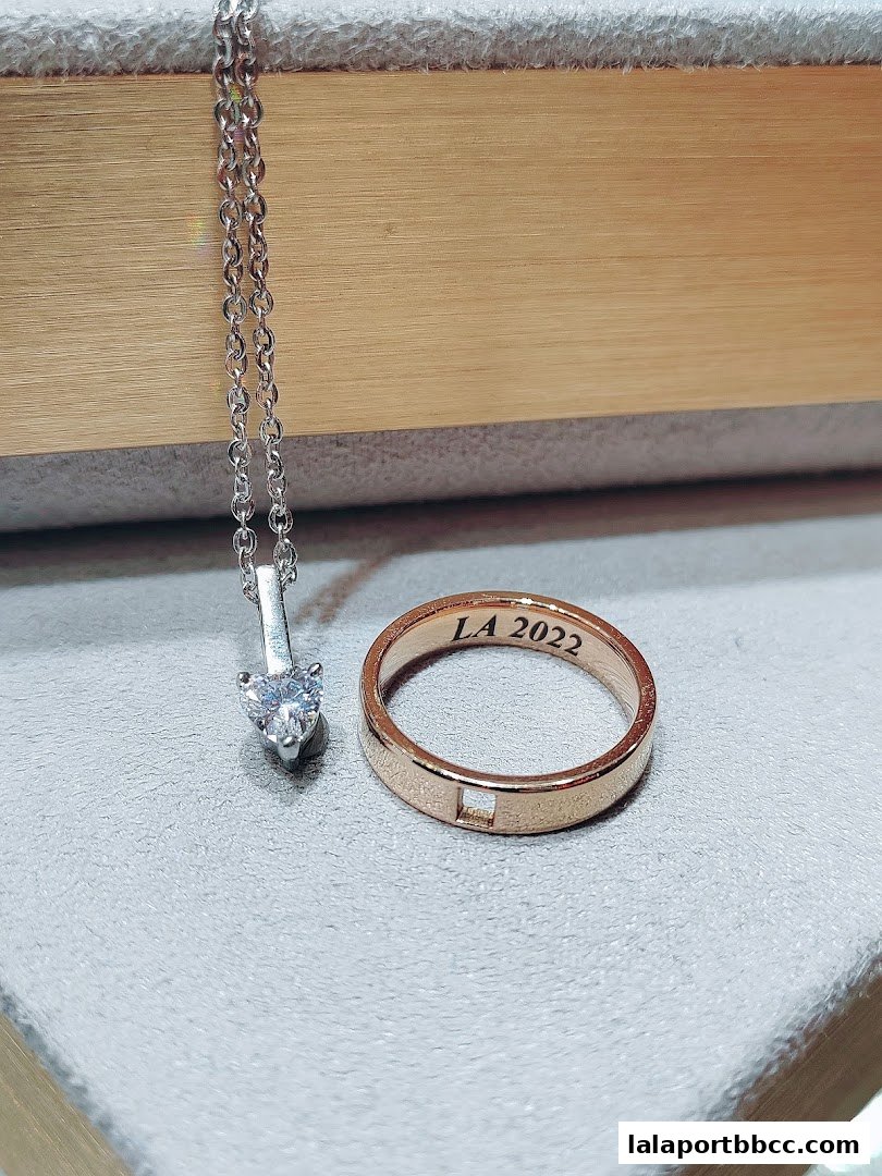 A close-up of a silver necklace with a heart-shaped diamond and a rose gold ring engraved with 'LA 2022'.