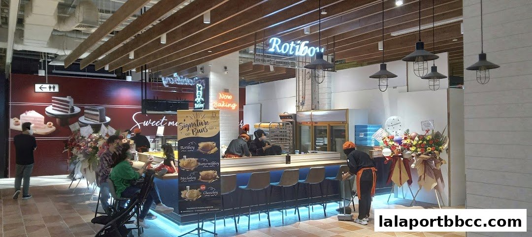Interior view of a bakery with customers and staff, showcasing baked goods and a 'Now Baking' sign.