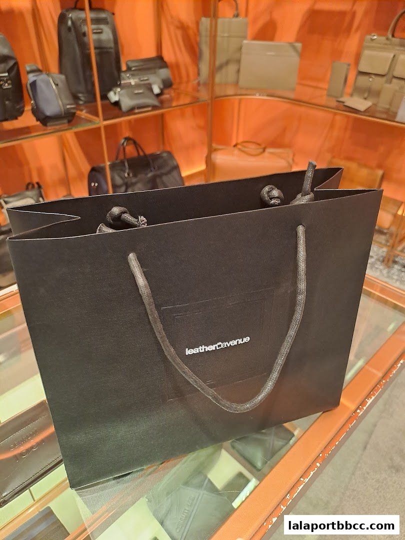 A black shopping bag with the 'Leather Avenue' logo sits on a glass surface, with bags displayed in the background.