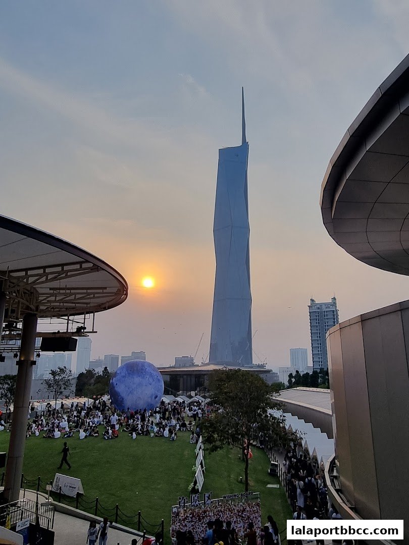 A crowd gathers on a grassy area with a large blue sphere and a tall building in the background at sunset.