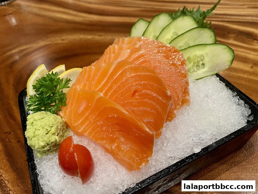 Fresh salmon sashimi served with wasabi, lemon, cucumber slices, and a cherry tomato.