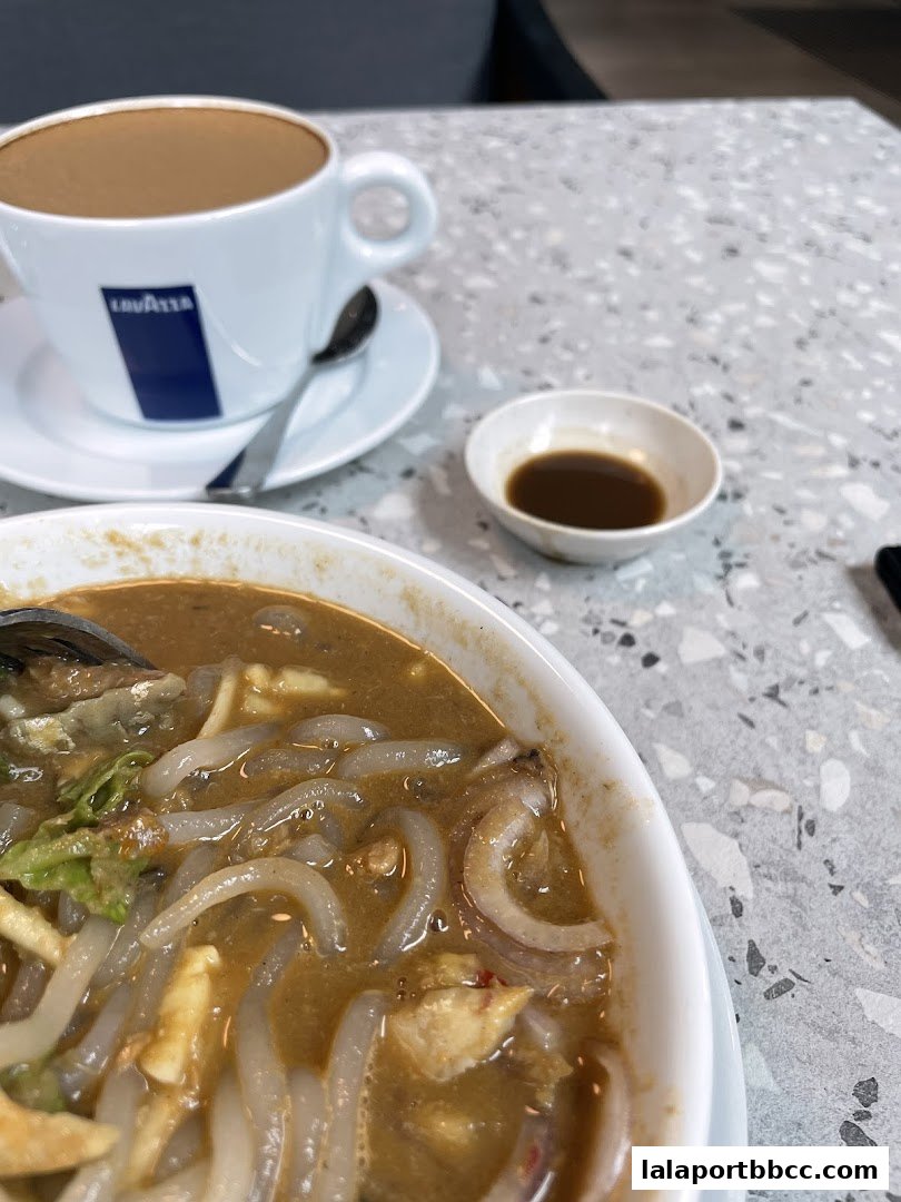 A close-up of a bowl of Laksa with a cup of coffee and a small dish of dipping sauce.