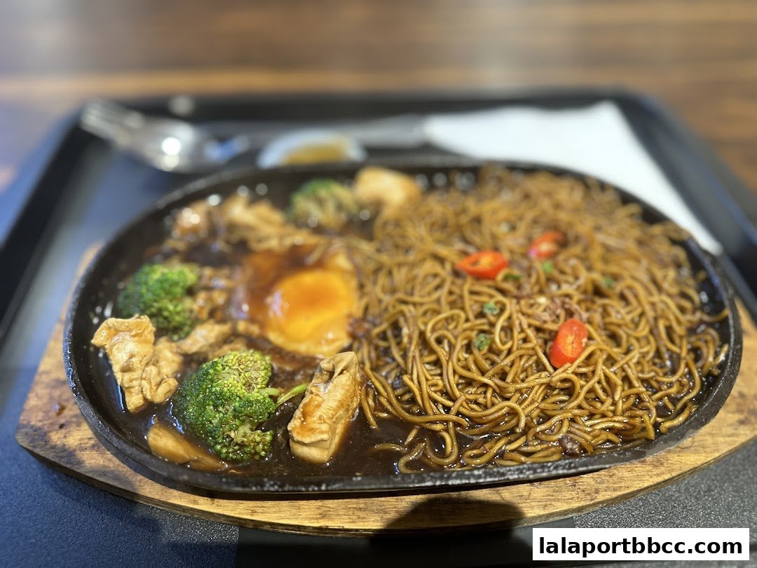 A sizzling plate of noodles with chicken, broccoli, and a fried egg.