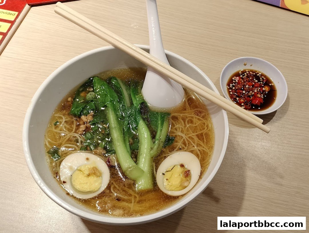 A bowl of steaming noodle soup with vegetables, egg, and a side of chili sauce.