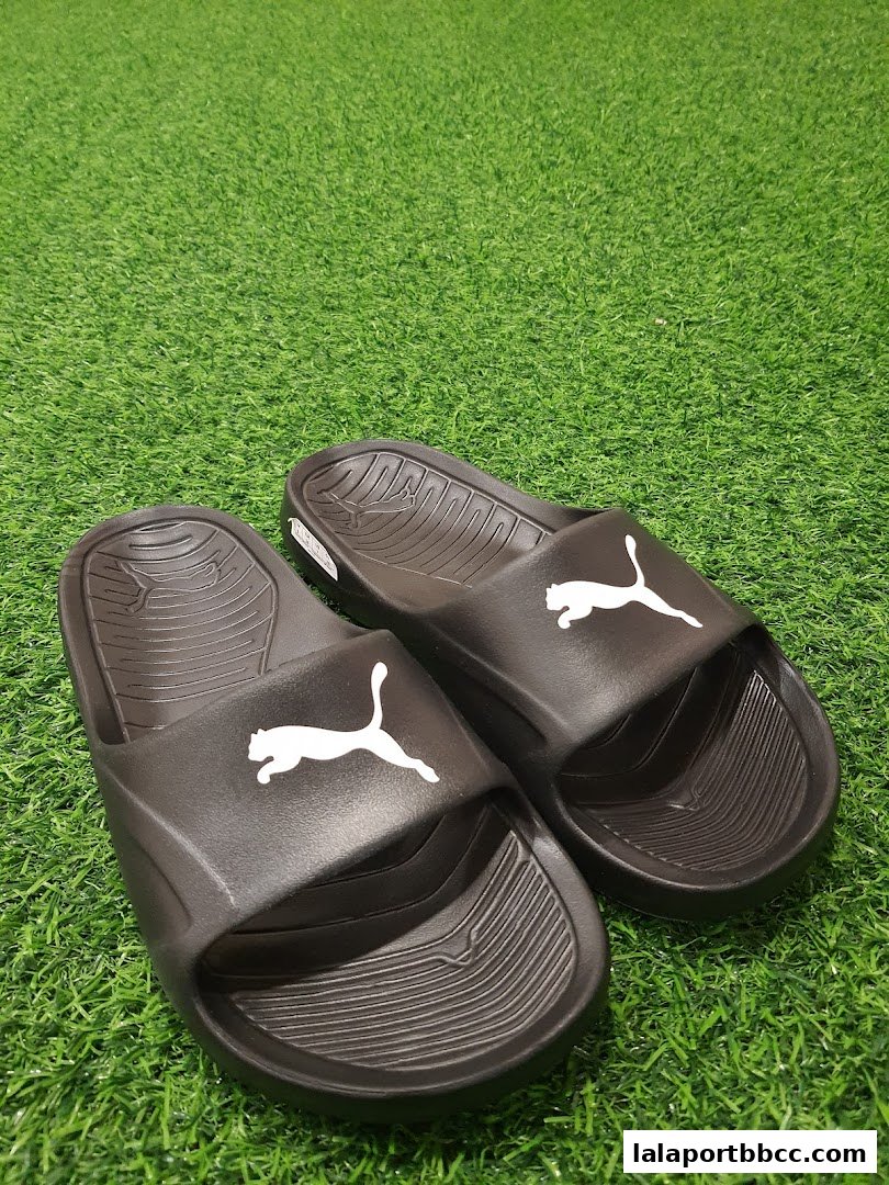 A pair of black Puma slide sandals with white Puma logos on a green grassy background.