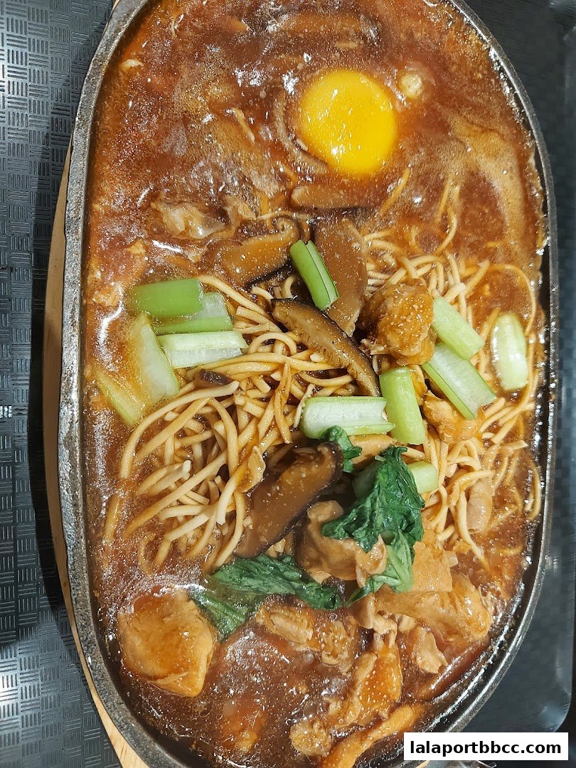 A sizzling claypot dish with noodles, egg, mushrooms, and vegetables in a rich sauce.