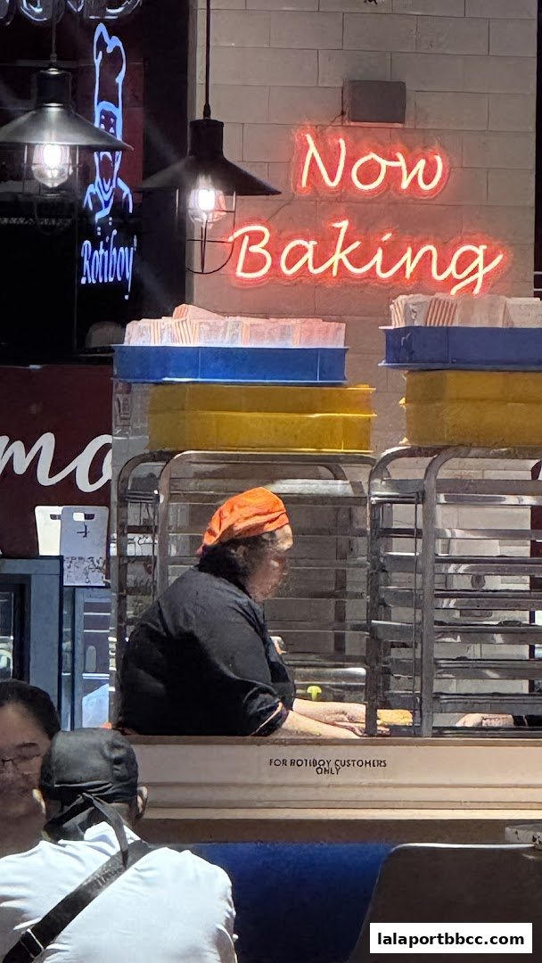 A baker wearing an orange headscarf works behind a counter with a neon sign saying 'Now Baking'.