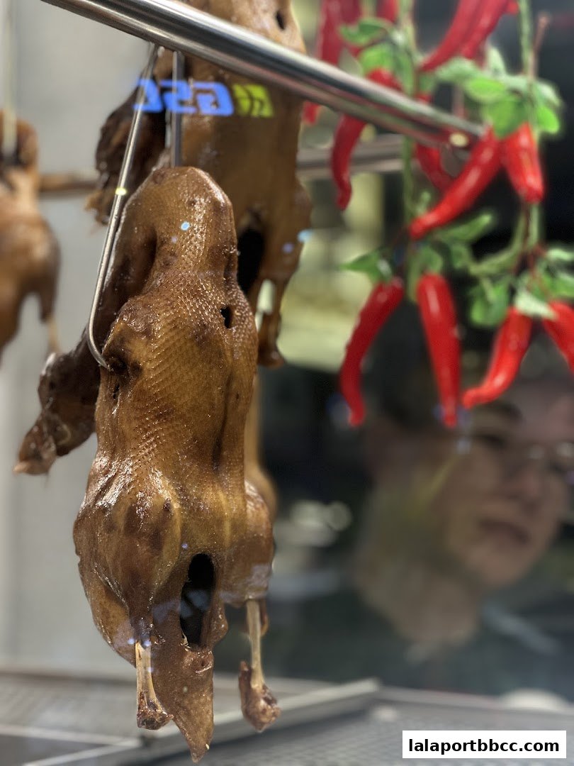 Close-up of roasted ducks hanging, with red chilies and a reflection of a person in the background.