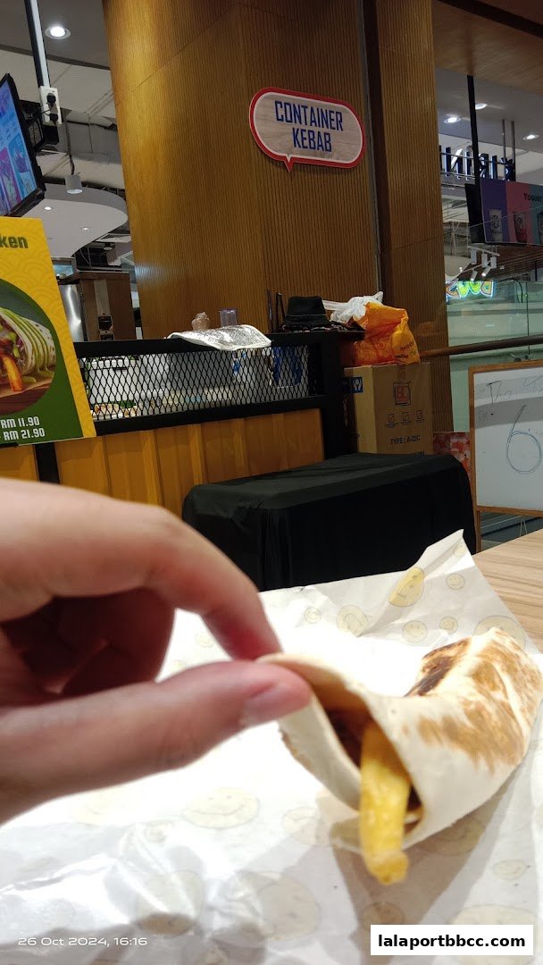 A hand holds a kebab wrap with fries peeking out, with the shop sign visible.