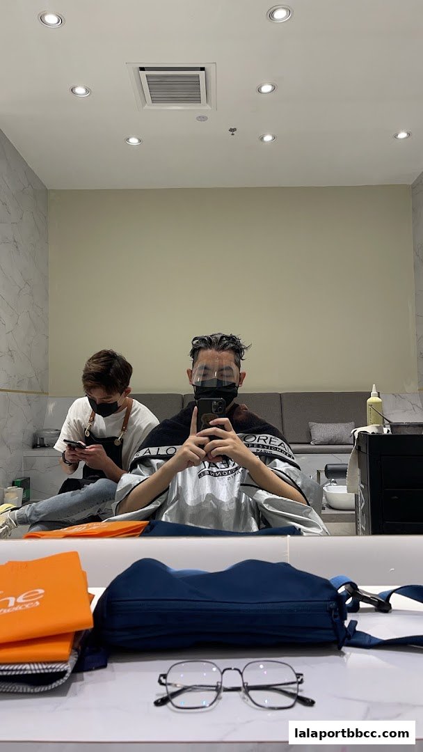 A person takes a mirror selfie in a salon with a stylist in the background.