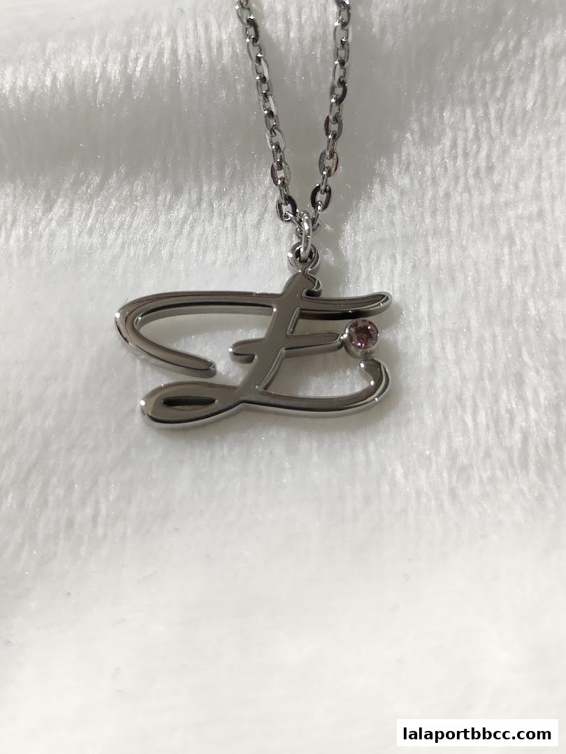 A close-up shot of a silver necklace with a stylized letter 'F' pendant and a small pink stone.