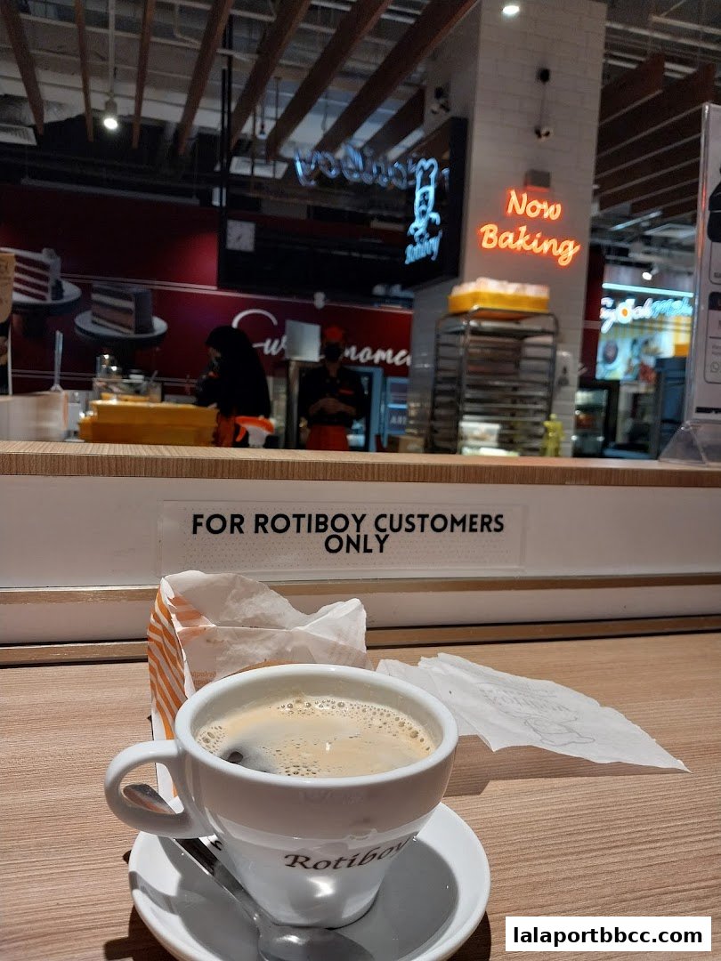 A cup of coffee and a bag of pastries from Rotiboy with a 'Now Baking' sign in the background.