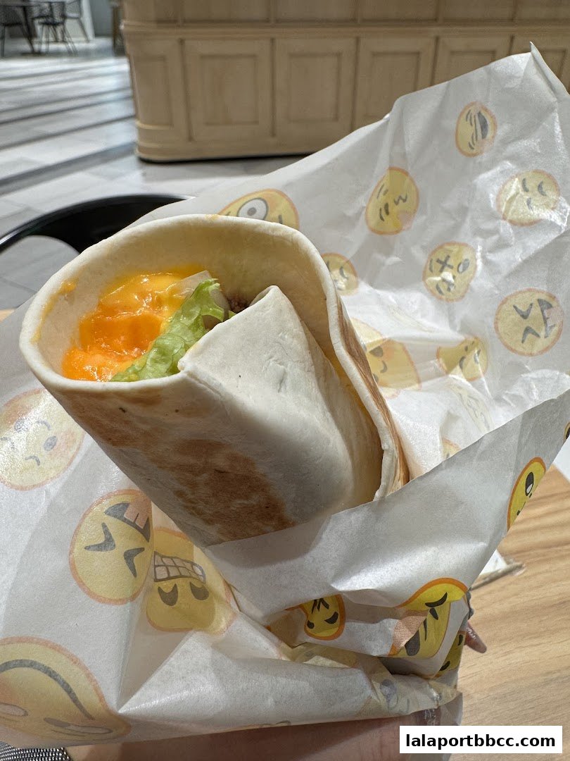 A close-up of a kebab wrap with lettuce and melted cheese, wrapped in emoji-patterned paper.
