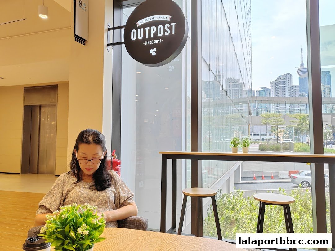 A woman sits at a table in a cafe, looking at her phone, with a city view outside.