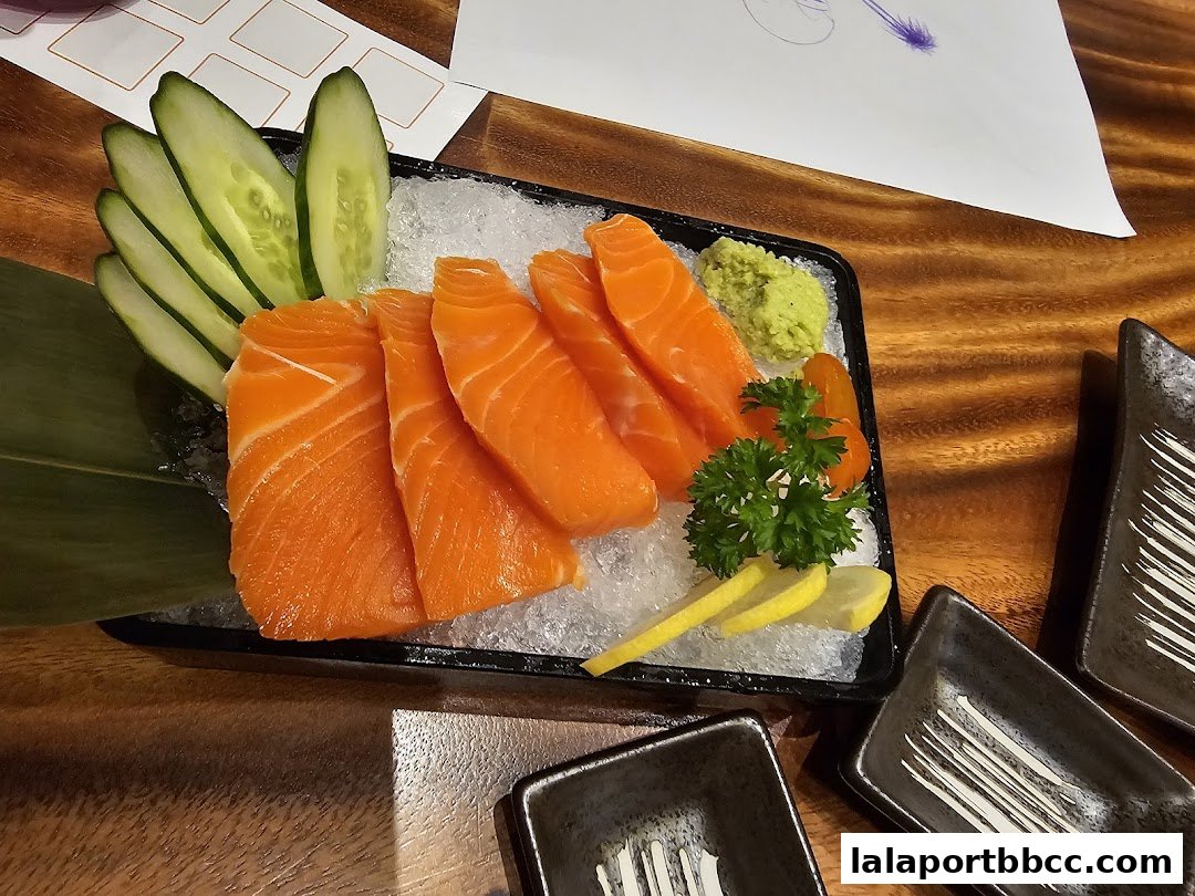 A plate of fresh salmon sashimi with cucumber slices, wasabi, and lemon wedges.