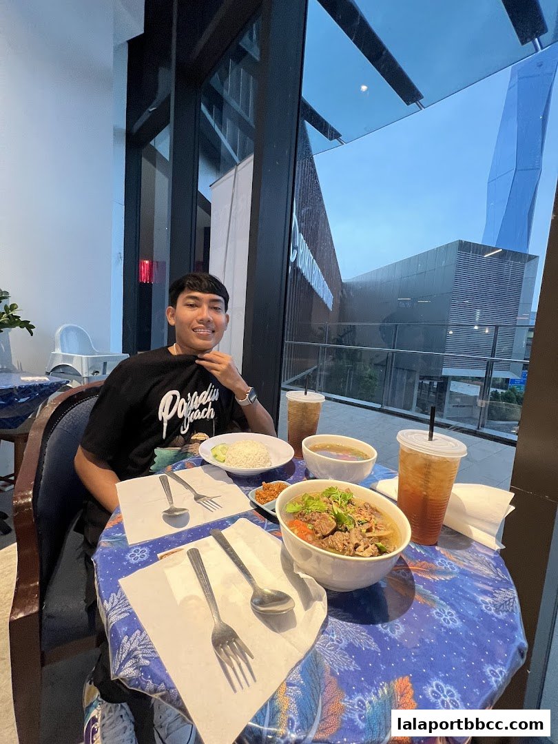 A man sits at a table with a meal and drinks, with a view of a modern building.