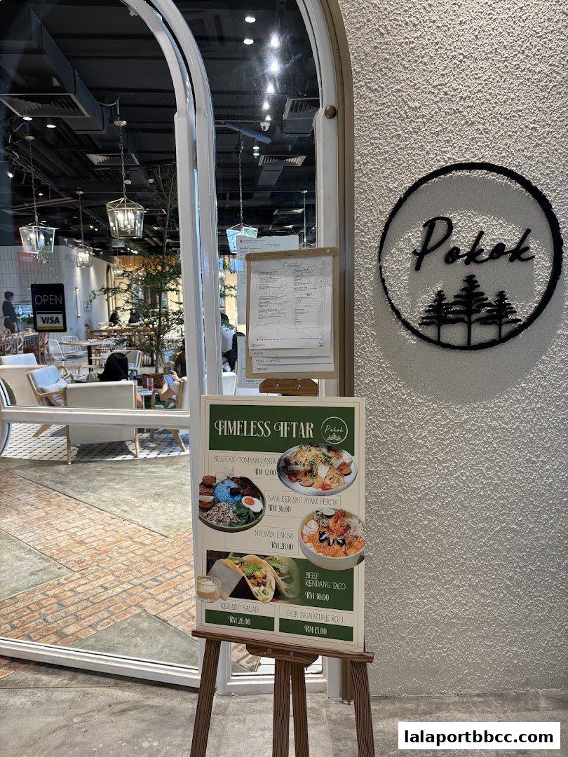 A sign outside POKOK LaLaport displays a menu with various Malaysian dishes and prices.