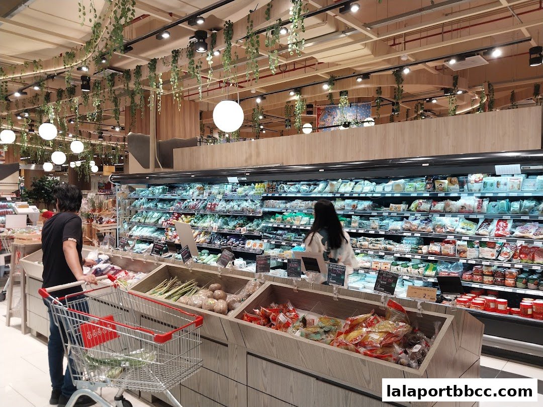 Shoppers browse fresh produce and packaged goods in a well-lit grocery store with hanging plants.