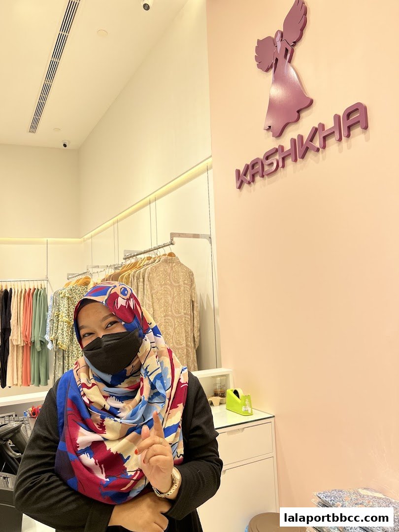 A woman in a colorful hijab smiles in a clothing store with the Kashkha logo.