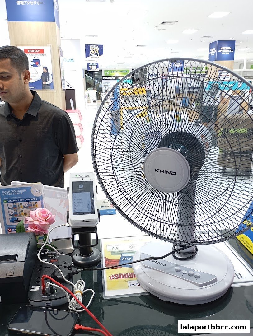 A man stands near a fan and payment terminal in a brightly lit electronics store.