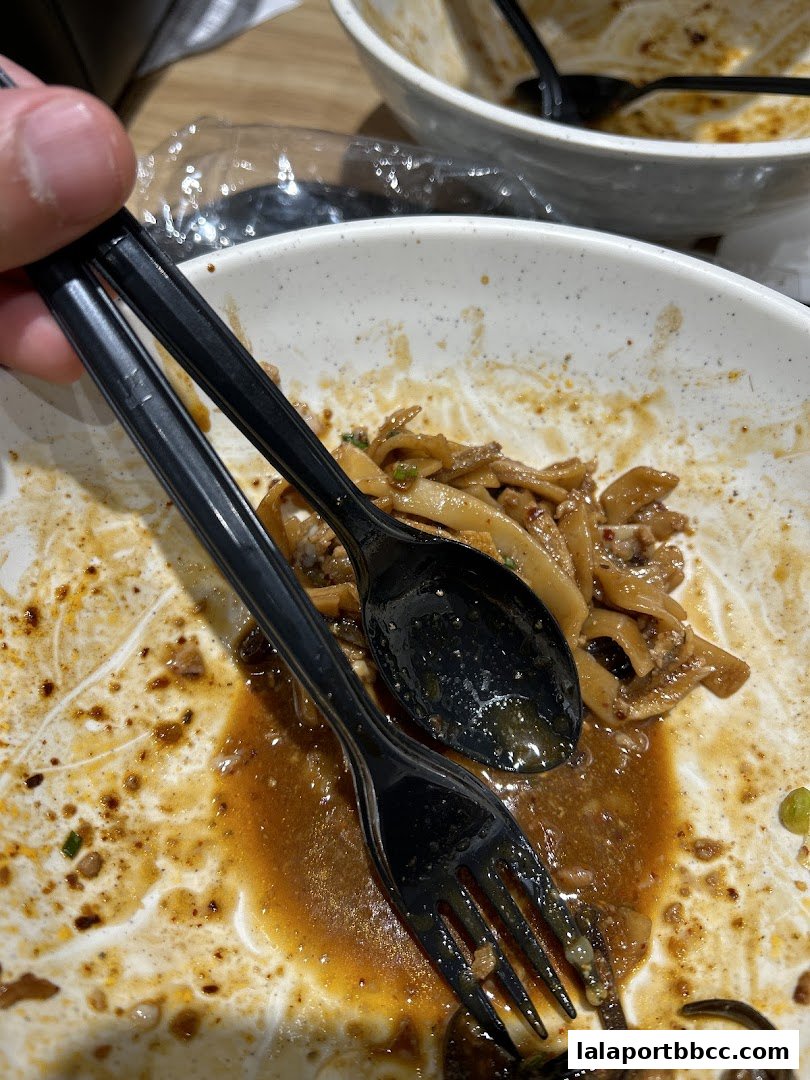 A close-up of a nearly empty bowl of noodles with a fork and spoon.