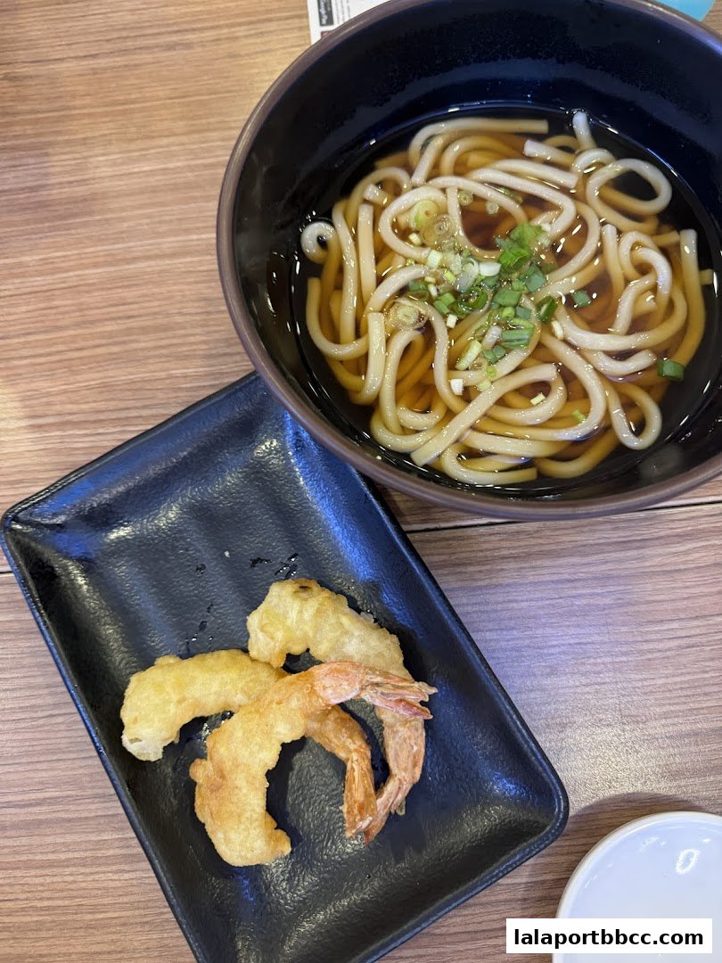 A bowl of udon noodles with tempura shrimp and vegetables on a black plate.
