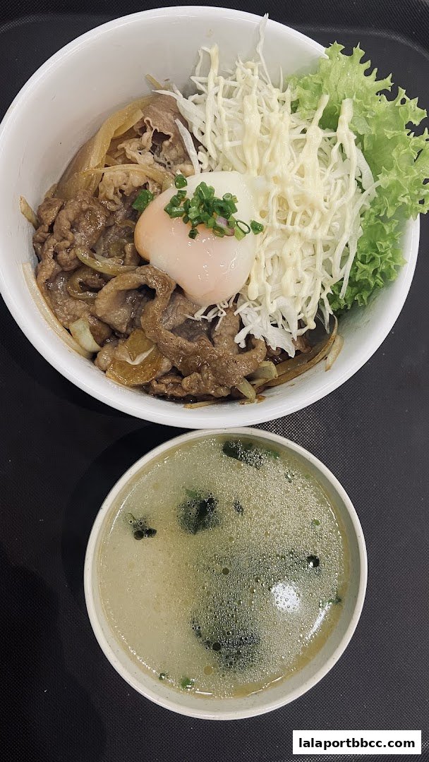 A bowl of Japanese curry rice with beef, onions, a soft-boiled egg, and shredded cabbage, served with a side of soup.