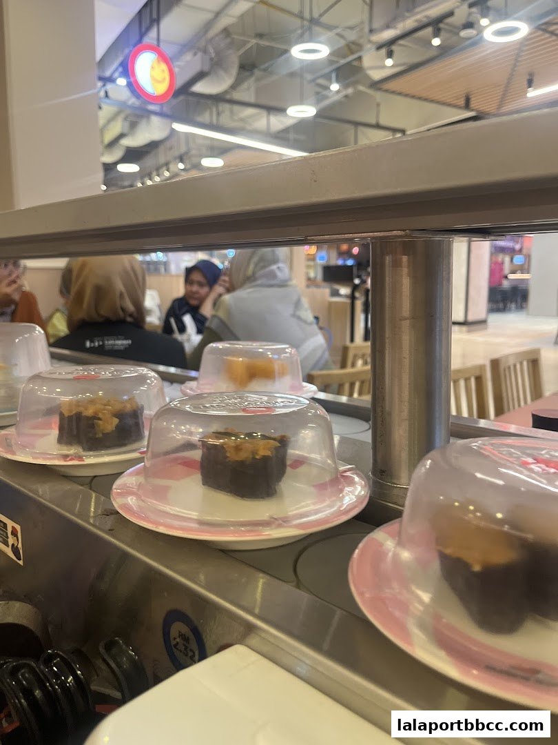 Sushi plates on a conveyor belt at Sushi King Lalaport BBCC with diners in the background.