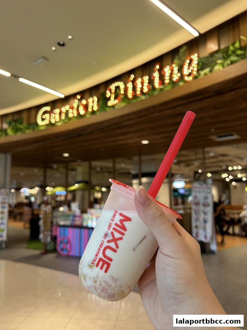 A hand holds a MIXUE ice cream tea with a red straw in front of a blurred mall background.