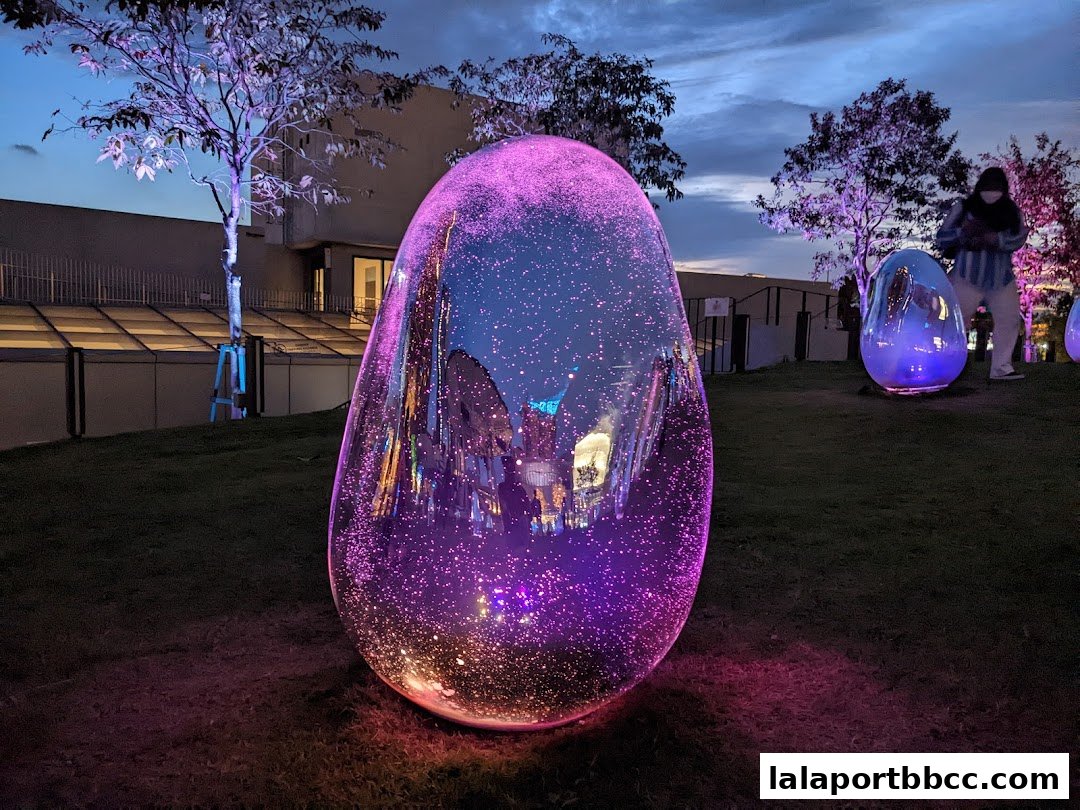 Large, glowing egg-shaped sculptures illuminated with purple lights on a grassy lawn at dusk.