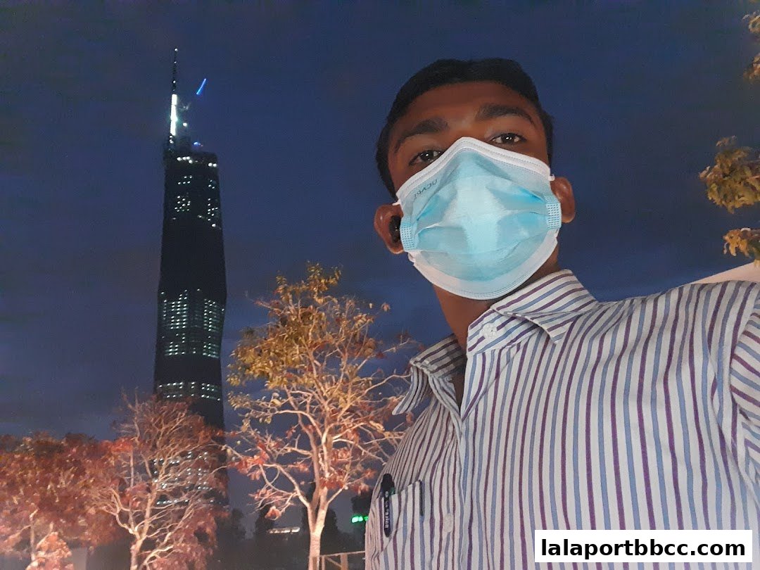 A person wearing a mask poses for a selfie with a tall skyscraper in the background at night.