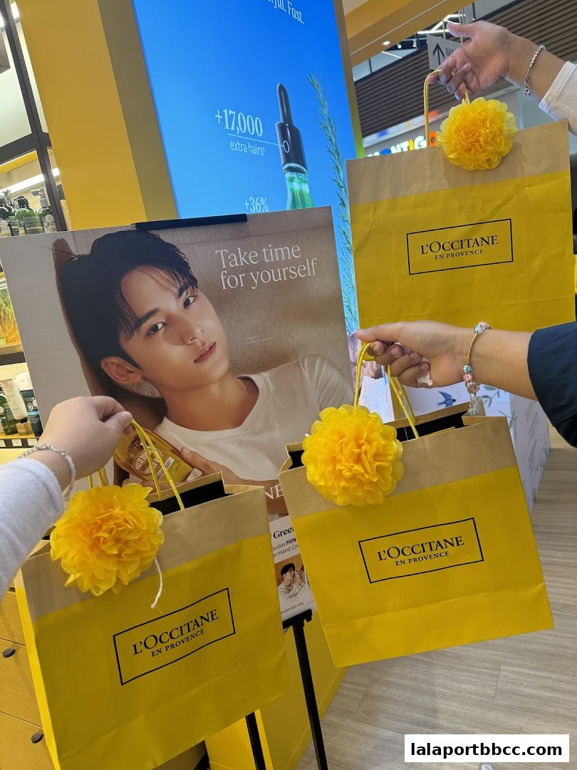 Hands holding L'Occitane shopping bags with yellow pom-poms and a promotional poster.