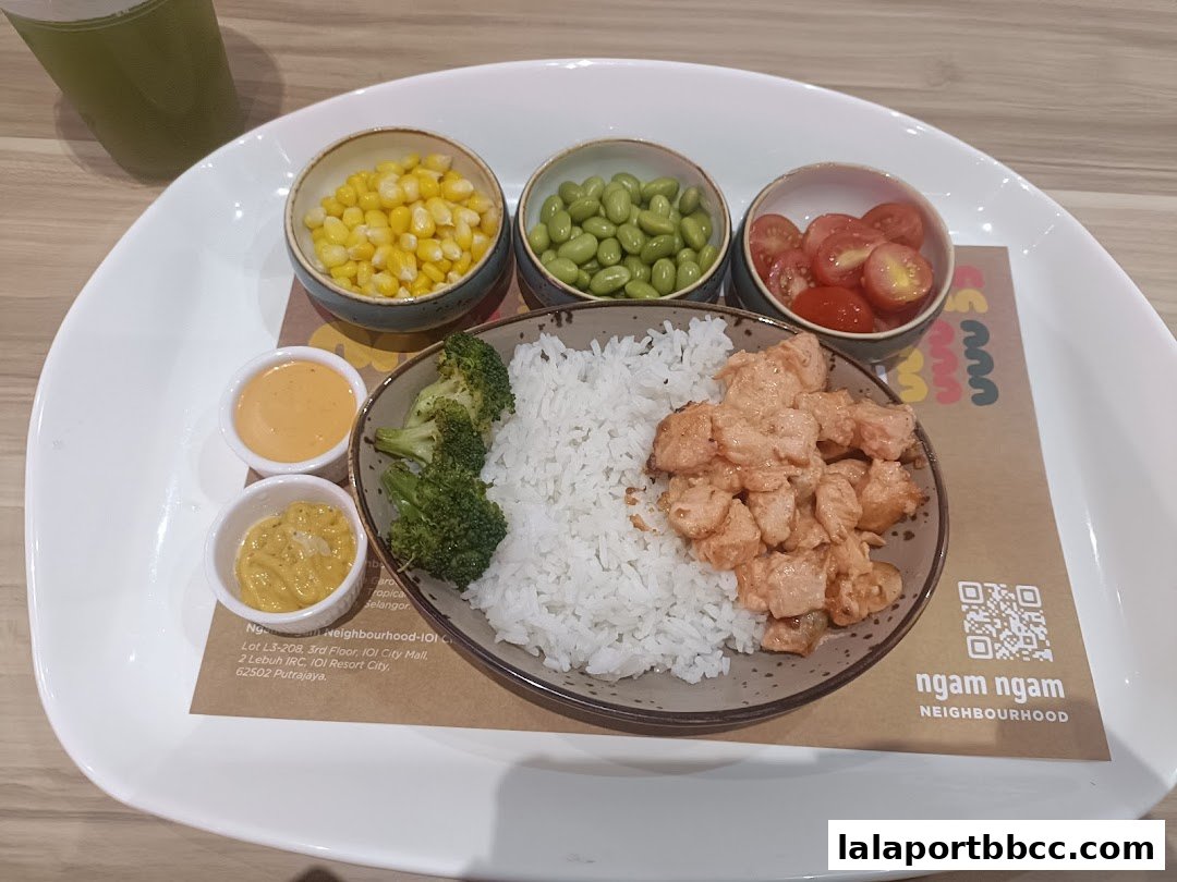 A plate of rice with salmon, broccoli, corn, edamame, and cherry tomatoes, served with two dipping sauces.