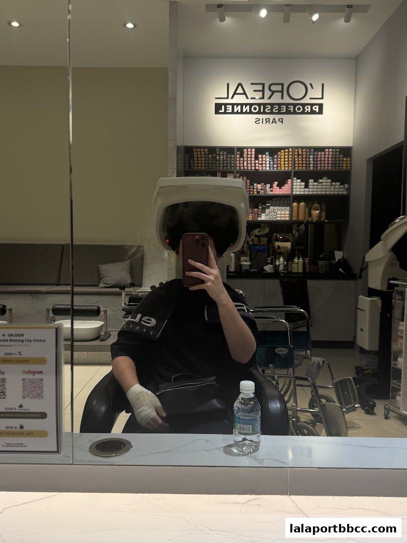 A person sits in a salon chair under a hair dryer, reflecting in a mirror.