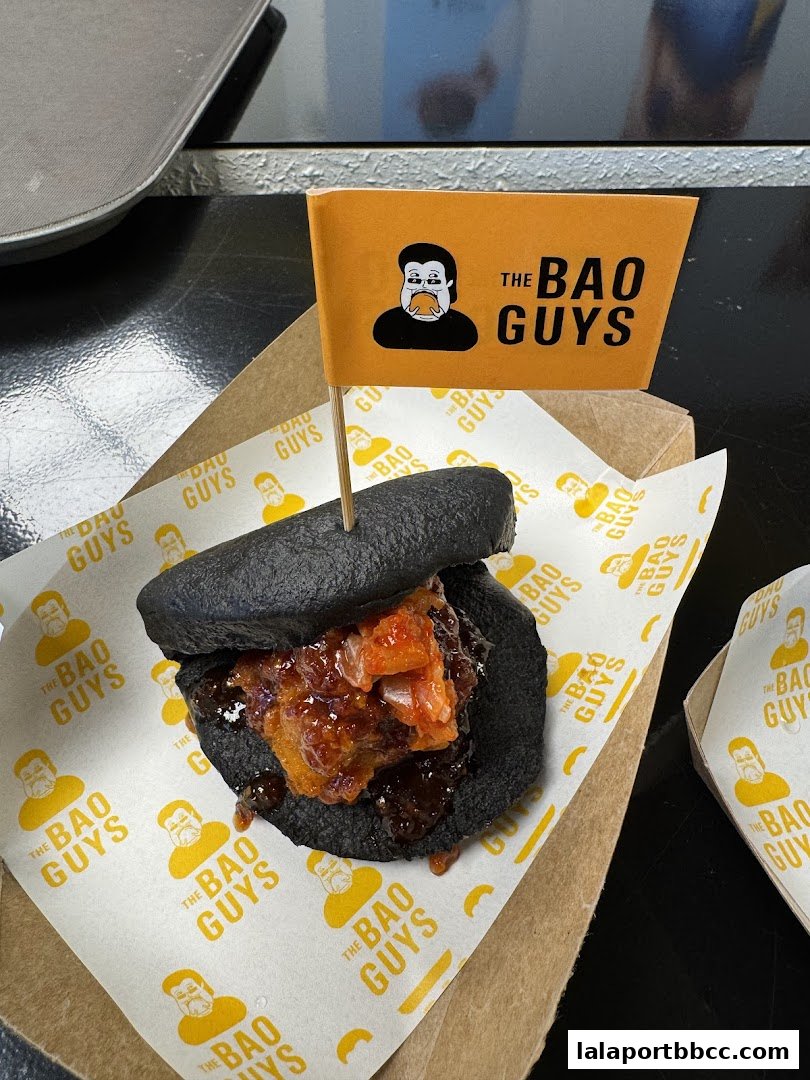 A black bao bun filled with savory ingredients, branded with 'The Bao Guys' logo.