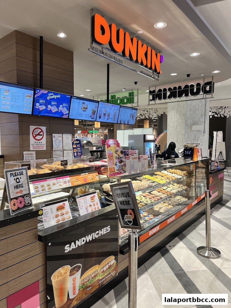A Dunkin' Donuts shop counter displaying donuts, sandwiches, and drinks with digital menus above.