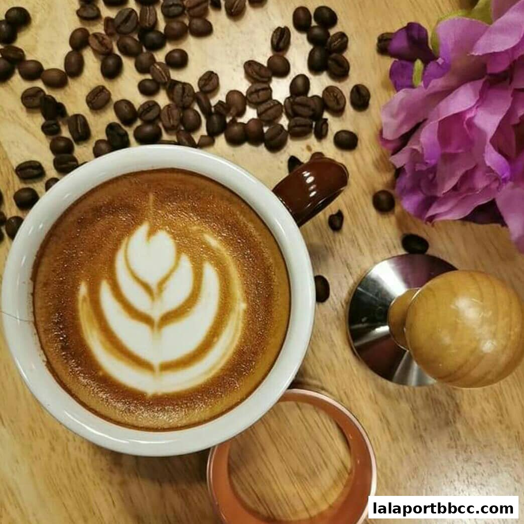 A close-up of a latte with intricate latte art, surrounded by coffee beans and a flower.