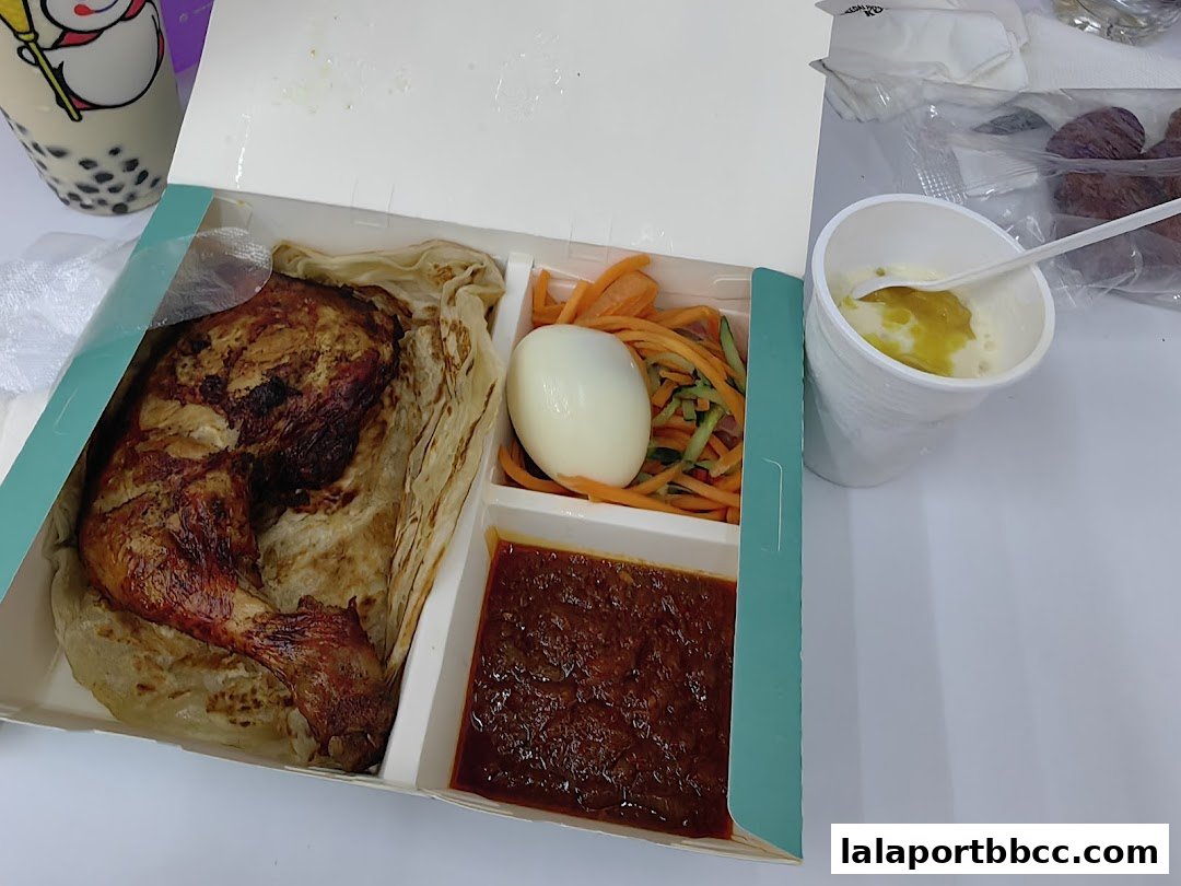 A takeaway box containing a roasted chicken leg with roti, salad, and a side of sauce, with a drink in the background.