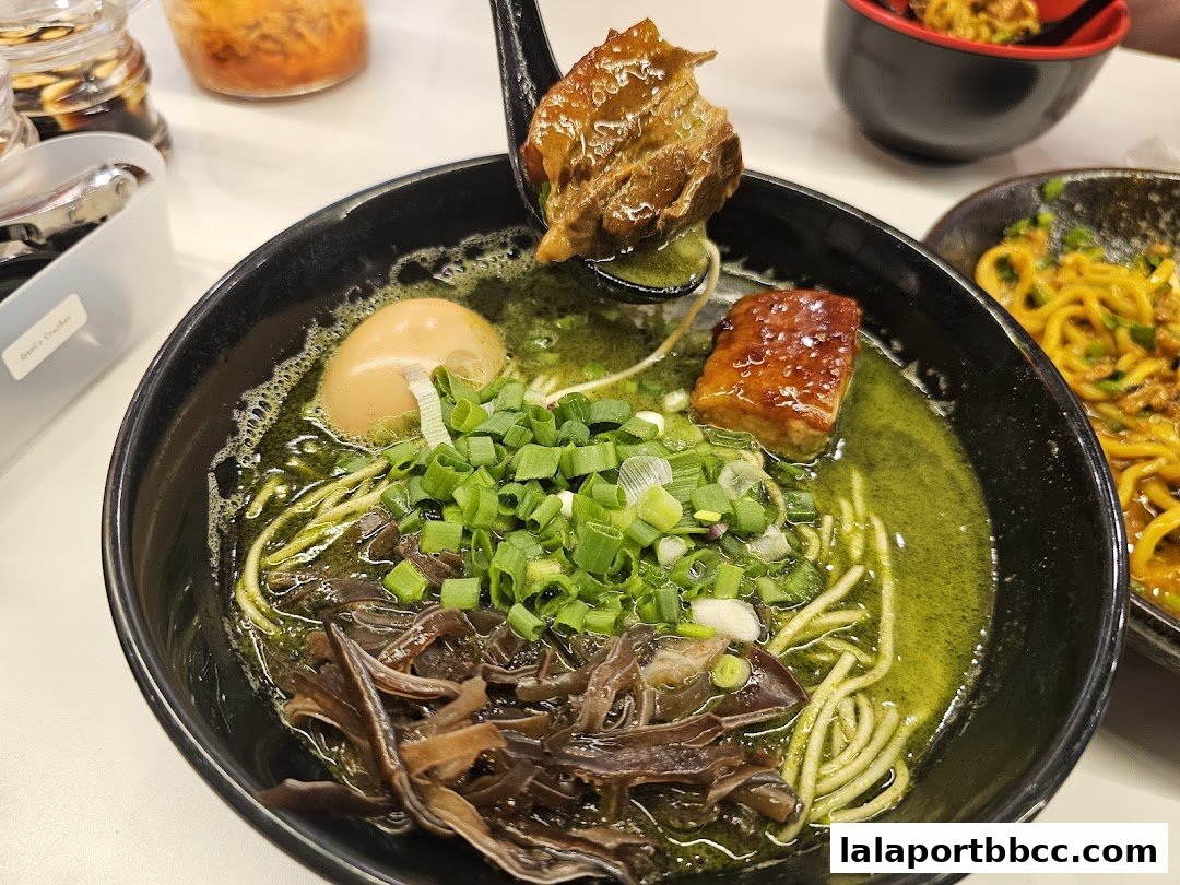 A close-up of a bowl of ramen with green broth, meat, egg, and green onions.