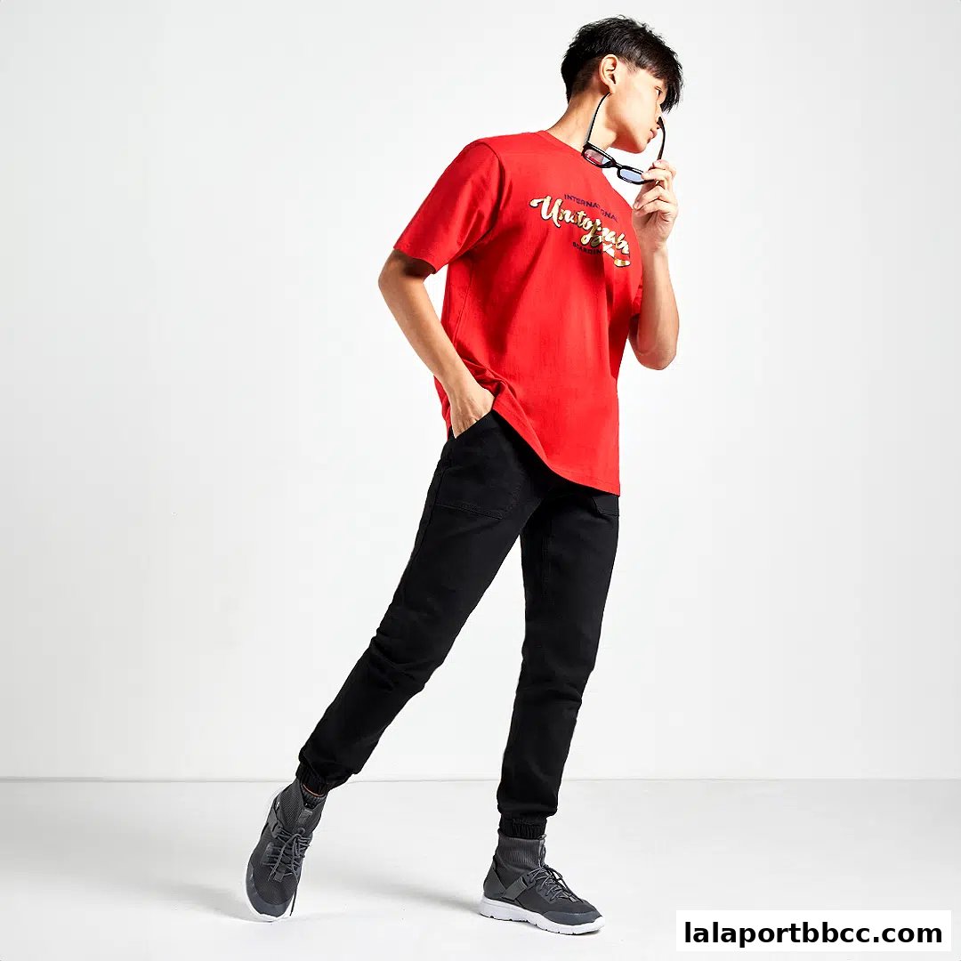 A young man models a red t-shirt and black pants, holding sunglasses.