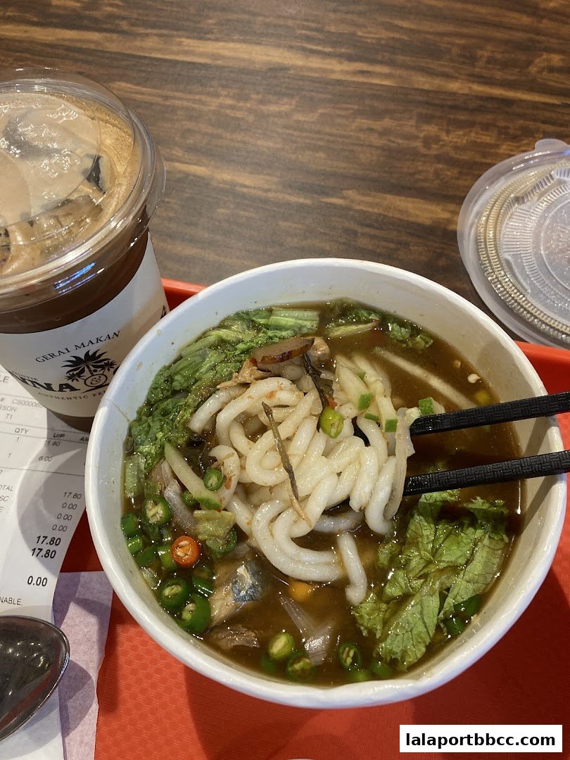 A bowl of noodles with broth, vegetables, and a drink on a wooden table.