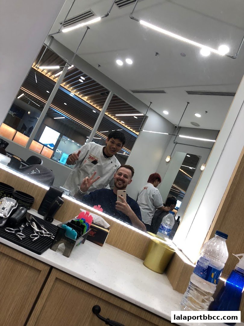 Two barbers and a customer smiling in a barbershop mirror, with styling tools visible.