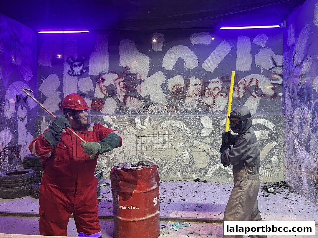 Two people in protective gear ready to smash a barrel with tools in a graffiti-covered room.