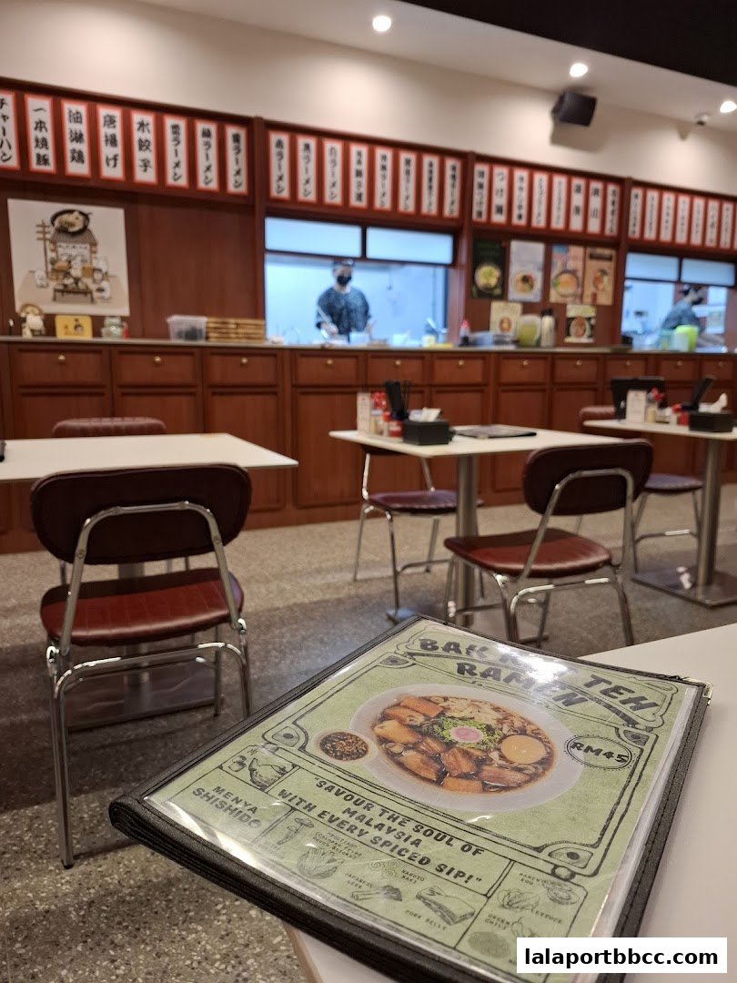 A menu for Bak Teh Ramen is placed on a table in a restaurant with a view of the kitchen.