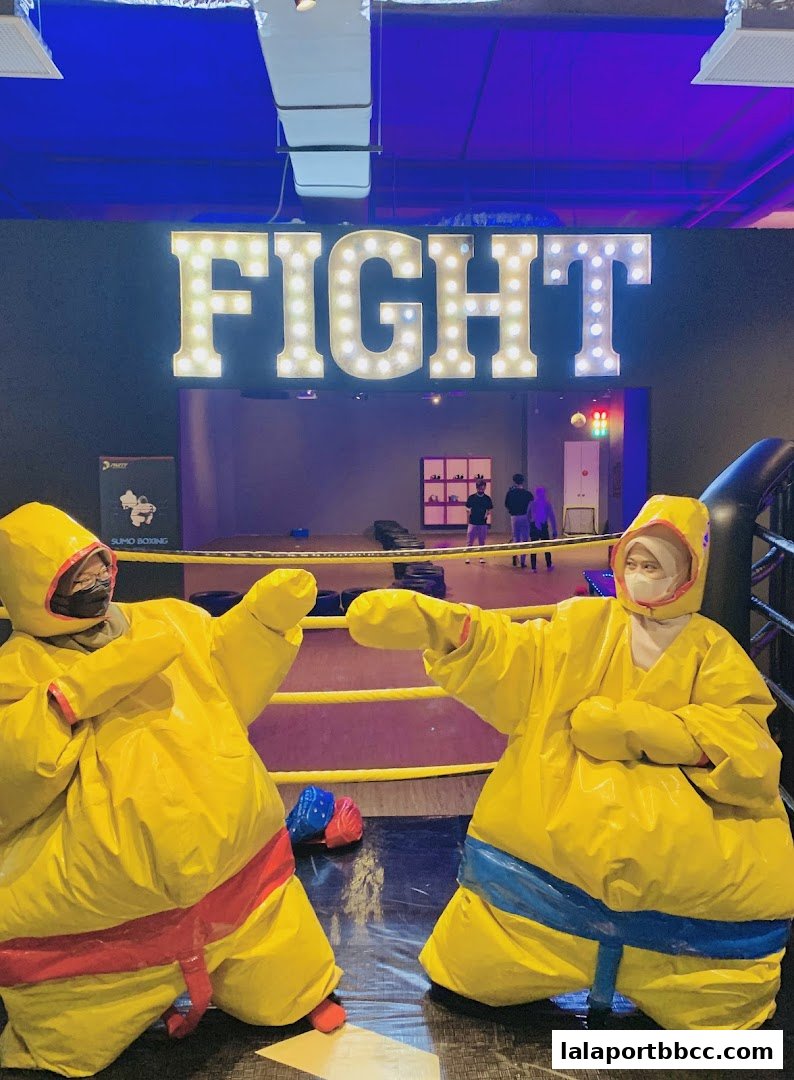 Two people in sumo wrestling suits in a boxing ring with a large 'FIGHT' sign.