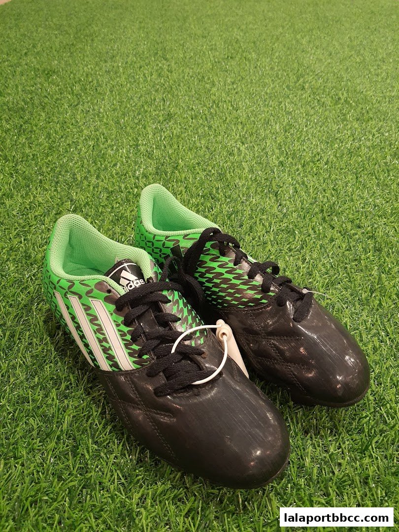 A pair of black and green Adidas soccer cleats are displayed on a green artificial turf surface.