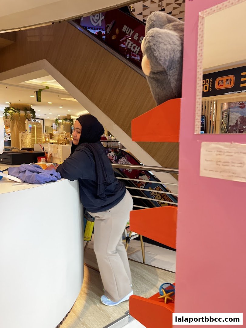 A woman leans over a counter in a brightly lit shopping mall, with escalators and displays visible.